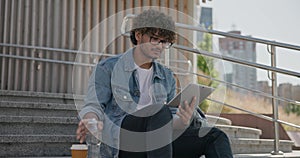 young handsome indian hindu man student freelancer sitting outdoors on steps using tablet pc computer pad