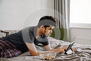 Young handsome indian man liying on bed working with tablet