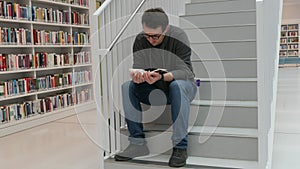 A young handsome intelligent man in glasses reads a book in a bookstore or library against the background of bookshelves