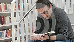 A young handsome intelligent man in glasses reads a book in a bookstore or library against the background of bookshelves