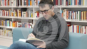 A young handsome intelligent man in glasses reads a book in a bookstore or library against the background of bookshelves