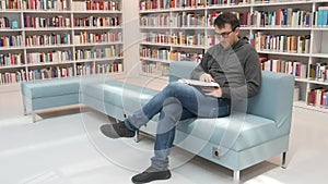A young handsome intelligent man in glasses reads a book in a bookstore or library against the background of bookshelves