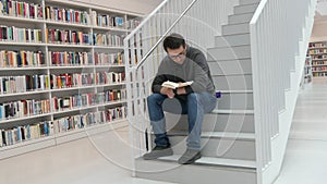 A young handsome intelligent man in glasses reads a book in a bookstore or library against the background of bookshelves