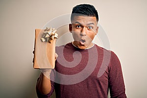 Young handsome latin man holding birthday gift over isolated white background scared in shock with a surprise face, afraid and