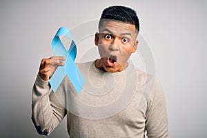 Young handsome latin man holding blue cancer ribbon over isolated white background scared in shock with a surprise face, afraid