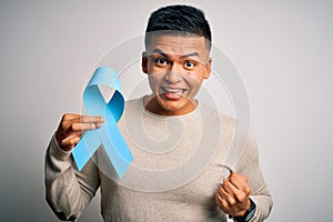 Young handsome latin man holding blue cancer ribbon over isolated white background screaming proud and celebrating victory and