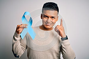 Young handsome latin man holding blue cancer ribbon over isolated white background surprised with an idea or question pointing