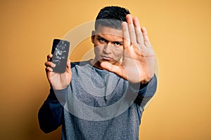 Young handsome latin man holding broken smartphone showing craked screen with open hand doing stop sign with serious and confident