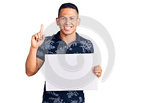 Young handsome latin man holding cardboard banner with blank space surprised with an idea or question pointing finger with happy