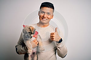 Young handsome latin man holding cute puppy pet over isolated white background happy with big smile doing ok sign, thumb up with