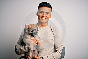 Young handsome latin man holding cute puppy pet over isolated white background with a happy face standing and smiling with a