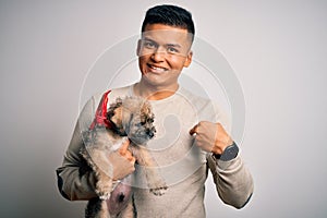 Young handsome latin man holding cute puppy pet over isolated white background with surprise face pointing finger to himself