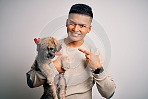 Young handsome latin man holding cute puppy pet over isolated white background very happy pointing with hand and finger