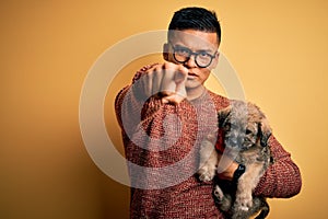 Young handsome latin man holding cute puppy pet over isolated yellow background pointing with finger to the camera and to you,