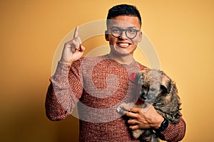 Young handsome latin man holding cute puppy pet over isolated yellow background surprised with an idea or question pointing finger