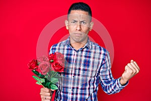 Young handsome latin man holding flowers annoyed and frustrated shouting with anger, yelling crazy with anger and hand raised