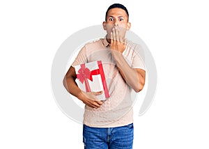 Young handsome latin man holding gift covering mouth with hand, shocked and afraid for mistake