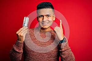 Young handsome latin man holding led lightbulb over isolated red background surprised with an idea or question pointing finger