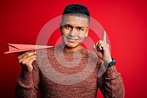 Young handsome latin man holding paper plane over isolated red background surprised with an idea or question pointing finger with