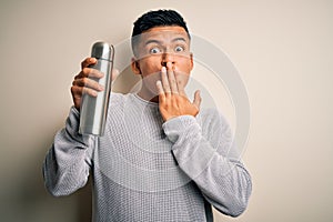 Young handsome latin man holding thermo with water over isolated white background cover mouth with hand shocked with shame for