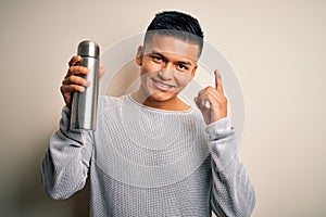 Young handsome latin man holding thermo with water over isolated white background surprised with an idea or question pointing