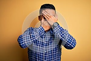 Young handsome latin man wearing casual shirt and glasses over yellow background Covering eyes and mouth with hands, surprised and