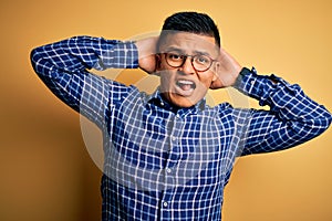 Young handsome latin man wearing casual shirt and glasses over yellow background Crazy and scared with hands on head, afraid and