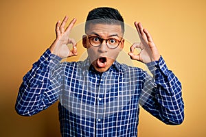 Young handsome latin man wearing casual shirt and glasses over yellow background looking surprised and shocked doing ok approval