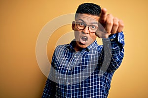 Young handsome latin man wearing casual shirt and glasses over yellow background Pointing with finger surprised ahead, open mouth