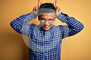 Young handsome latin man wearing casual shirt and glasses over yellow background Posing funny and crazy with fingers on head as