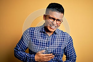 Young handsome latin man wearing casual shirt and glasses over yellow background smiling and laughing hard out loud because funny