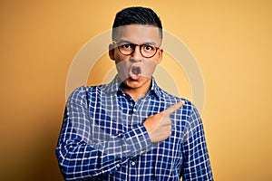 Young handsome latin man wearing casual shirt and glasses over yellow background Surprised pointing with finger to the side, open