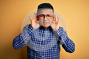 Young handsome latin man wearing casual shirt and glasses over yellow background Trying to hear both hands on ear gesture, curious