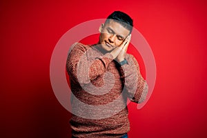 Young handsome latin man wearing casual sweater standing over red background sleeping tired dreaming and posing with hands