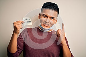 Young handsome latin man wearing medical mask holding paper with virus alert message surprised with an idea or question pointing