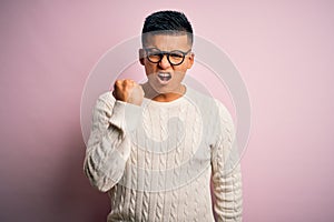 Young handsome latin man wearing white casual sweater and glasses over pink background angry and mad raising fist frustrated and
