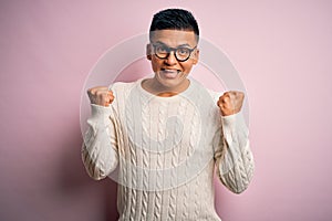 Young handsome latin man wearing white casual sweater and glasses over pink background celebrating surprised and amazed for