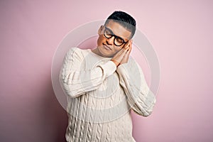 Young handsome latin man wearing white casual sweater and glasses over pink background sleeping tired dreaming and posing with
