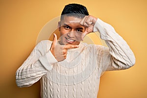 Young handsome latin man wearing white casual sweater over yellow background smiling making frame with hands and fingers with
