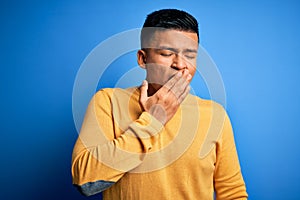 Young handsome latin man wearing yellow casual sweater over isolated blue background bored yawning tired covering mouth with hand