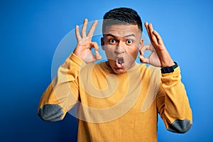 Young handsome latin man wearing yellow casual sweater over isolated blue background looking surprised and shocked doing ok