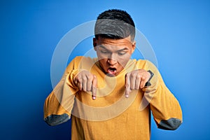 Young handsome latin man wearing yellow casual sweater over isolated blue background Pointing down with fingers showing