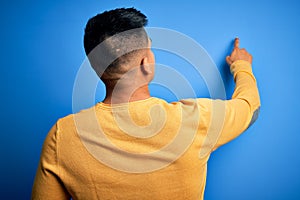 Young handsome latin man wearing yellow casual sweater over isolated blue background Posing backwards pointing ahead with finger