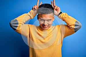 Young handsome latin man wearing yellow casual sweater over isolated blue background Posing funny and crazy with fingers on head