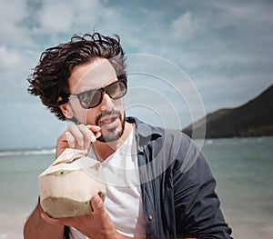 Young handsome latino man guy in sunglasses on the tropical shore of a tropical beach with fresh coconuts in his hands