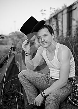 Young handsome mad, wearing grey pants and white t-shirt, holding black classic hat, sitting on abandoned railway track by old