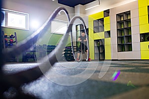 young and handsome male athlete man doing exercises for the muscles of the body with a battle ropes in fitness gym with