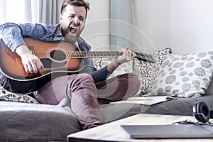 A young, handsome male composer plays an acoustic guitar and loudly sings a song of his own composition, sitting on a sofa, in a