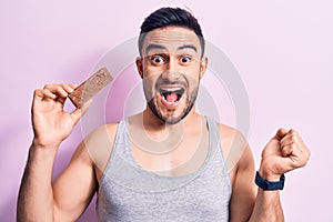 Young handsome man with beard eating energy protein bar over isolated pink background screaming proud, celebrating victory and