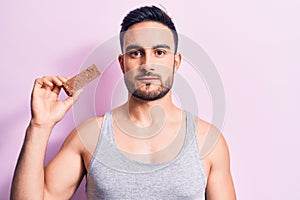 Young handsome man with beard eating energy protein bar over isolated pink background thinking attitude and sober expression photo